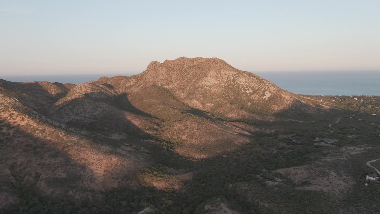 Third aerial frame from Cabo Pulmo showing the ridge shifted slightly right in the composition, with scrubby valley terrain below, the winding road visible at lower right, sea beyond, and no visible people.