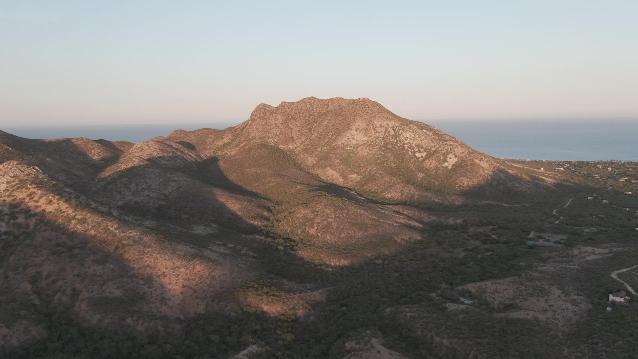 Mid-clip aerial view over Cabo Pulmo with more dark foreground terrain, a sunlit rocky ridge across the center, a thin winding road at lower right, distant sea, and no visible people.