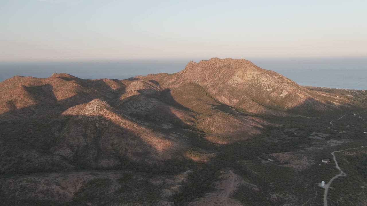 Cabo Pulmo aerial frame with a bright rocky ridge on the right, darker foreground hills, dry vegetation, a winding dirt road below, distant sea, and no visible people.