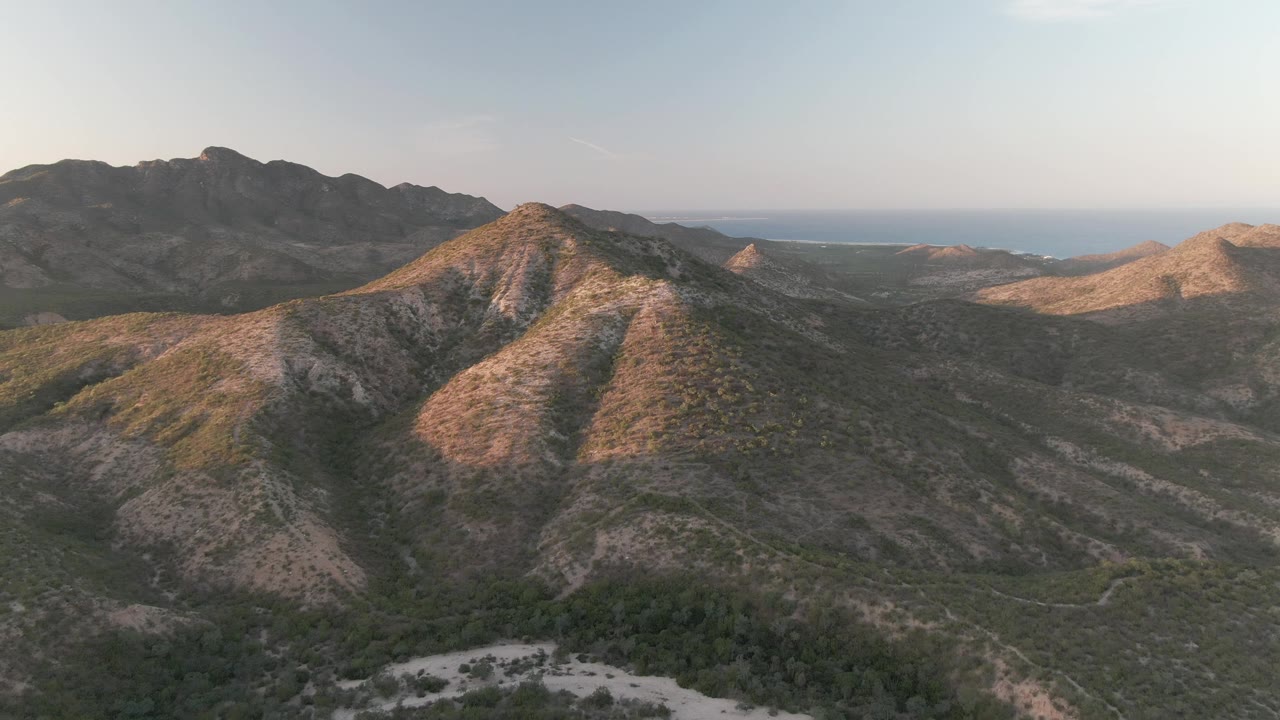 Aerial view over Cabo Pulmo mountain ridges and scrub-covered valley, with a pale path below, ocean on the far horizon, and no visible people.