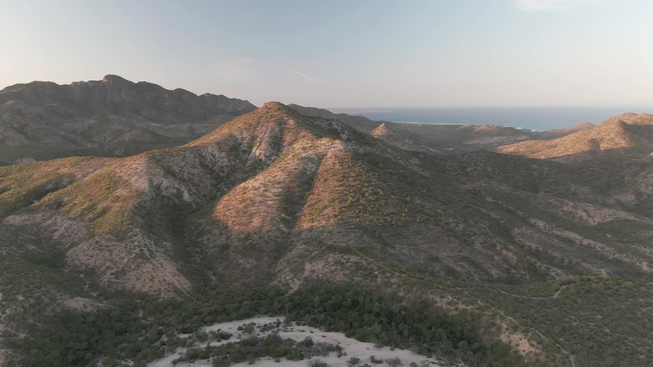 Wide Cabo Pulmo drone frame with a rounded sunlit hill centered over a green and tan valley, darker mountains behind, distant ocean, and no visible people.