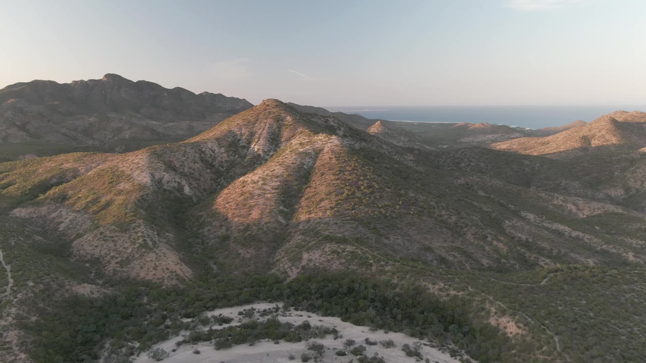 Cabo Pulmo aerial frame showing a pale winding path cutting through sparse valley vegetation, with layered hills, ocean haze at the horizon, and no visible people.