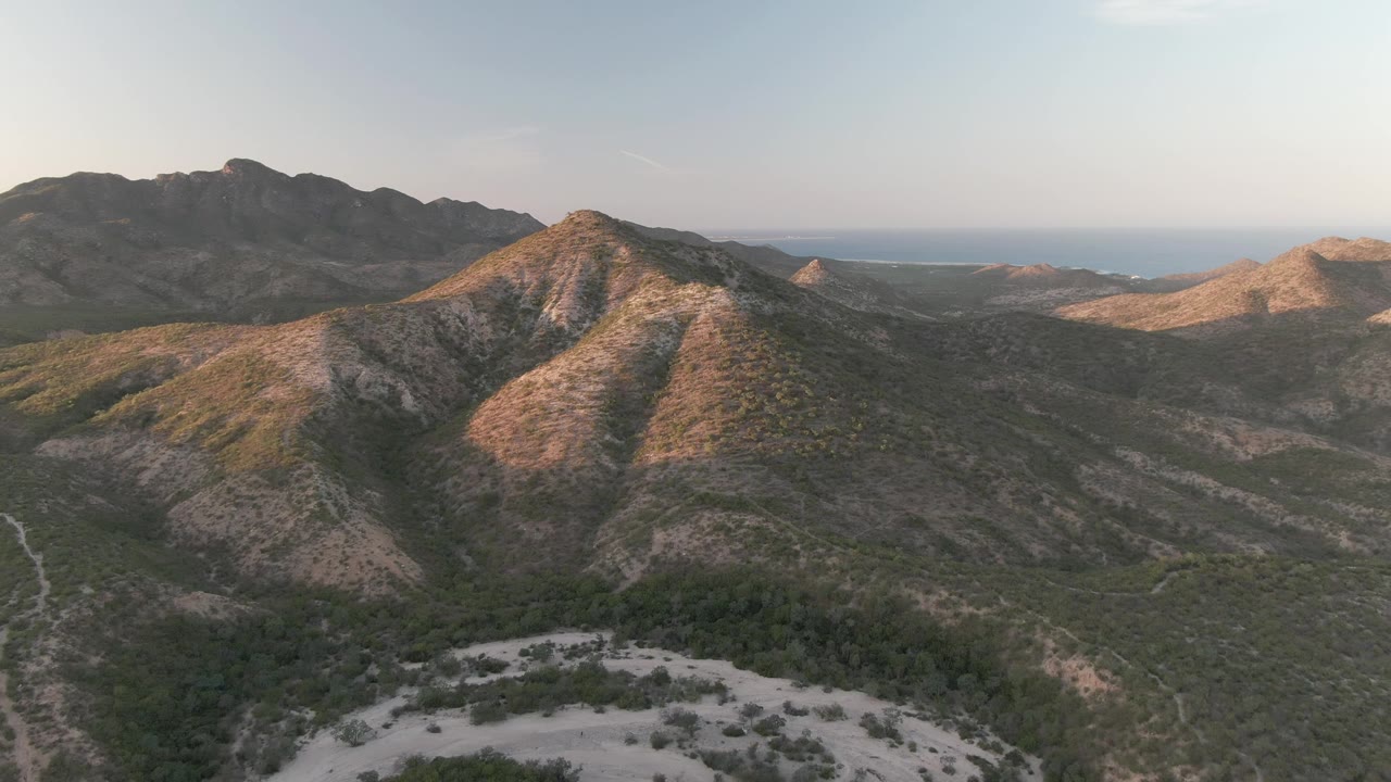 Aerial view in Cabo Pulmo with shaded rear ridges, a sunlit foreground slope, scrubland across the valley, distant sea, and no visible people.