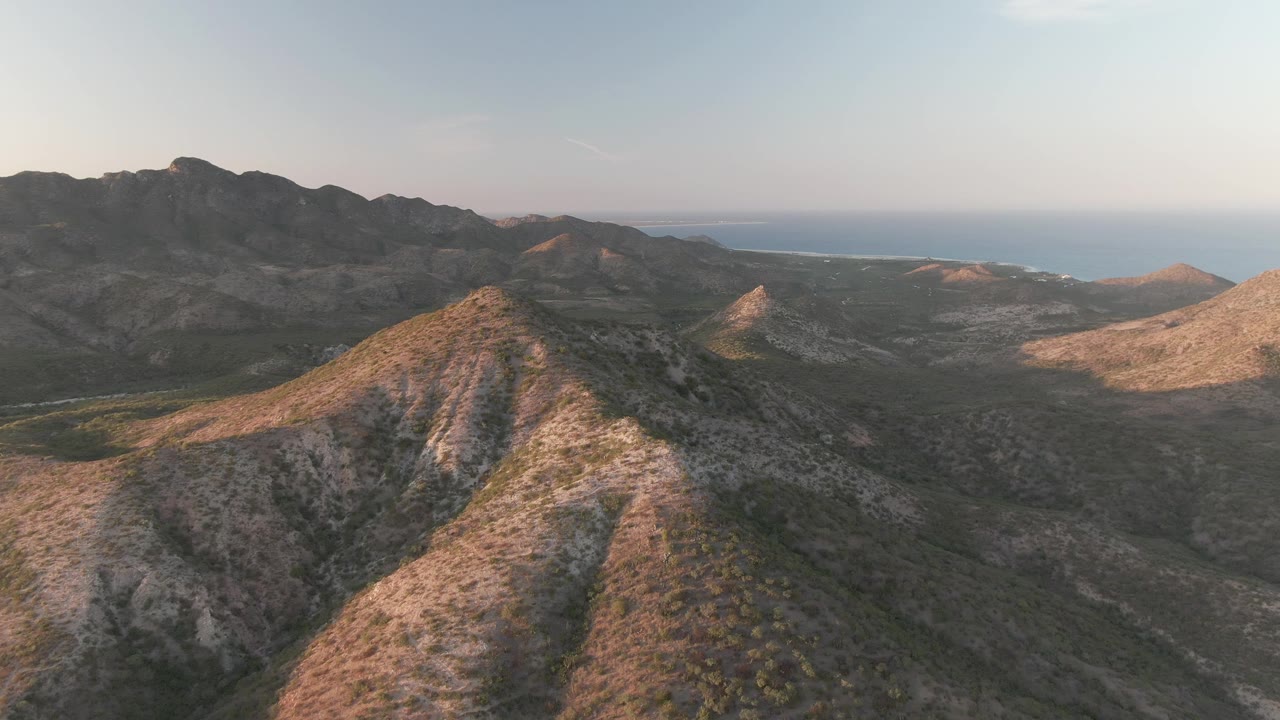 Aerial view over rocky Cabo Pulmo ridges with sparse greenery, darker mountains in the distance, a blue ocean horizon, and no visible people.