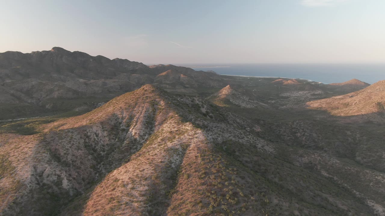 High aerial view of a Cabo Pulmo mountain slope, with tan rock faces, scattered shrubs, dark ridgelines behind, ocean on the horizon, and no visible people.
