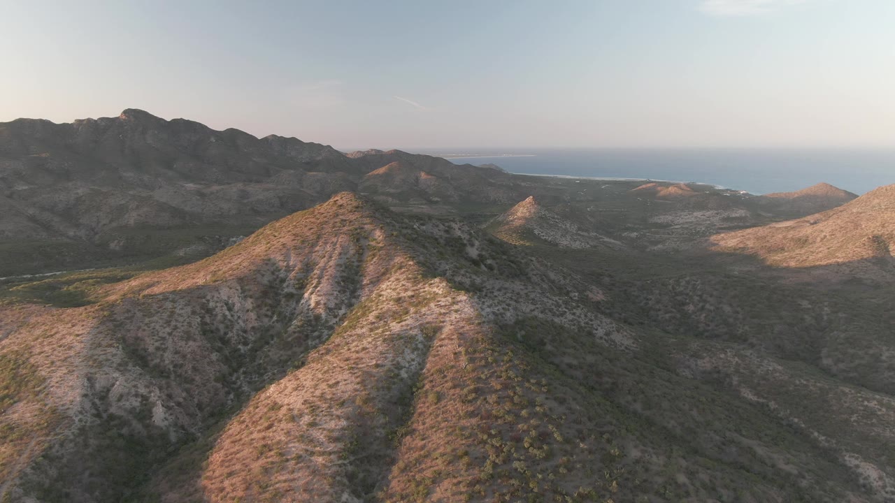 Cabo Pulmo ridge crest seen from above, with warm sunlight on the rocky peak, shadowed gullies below, distant mountains, ocean horizon, and no visible people.