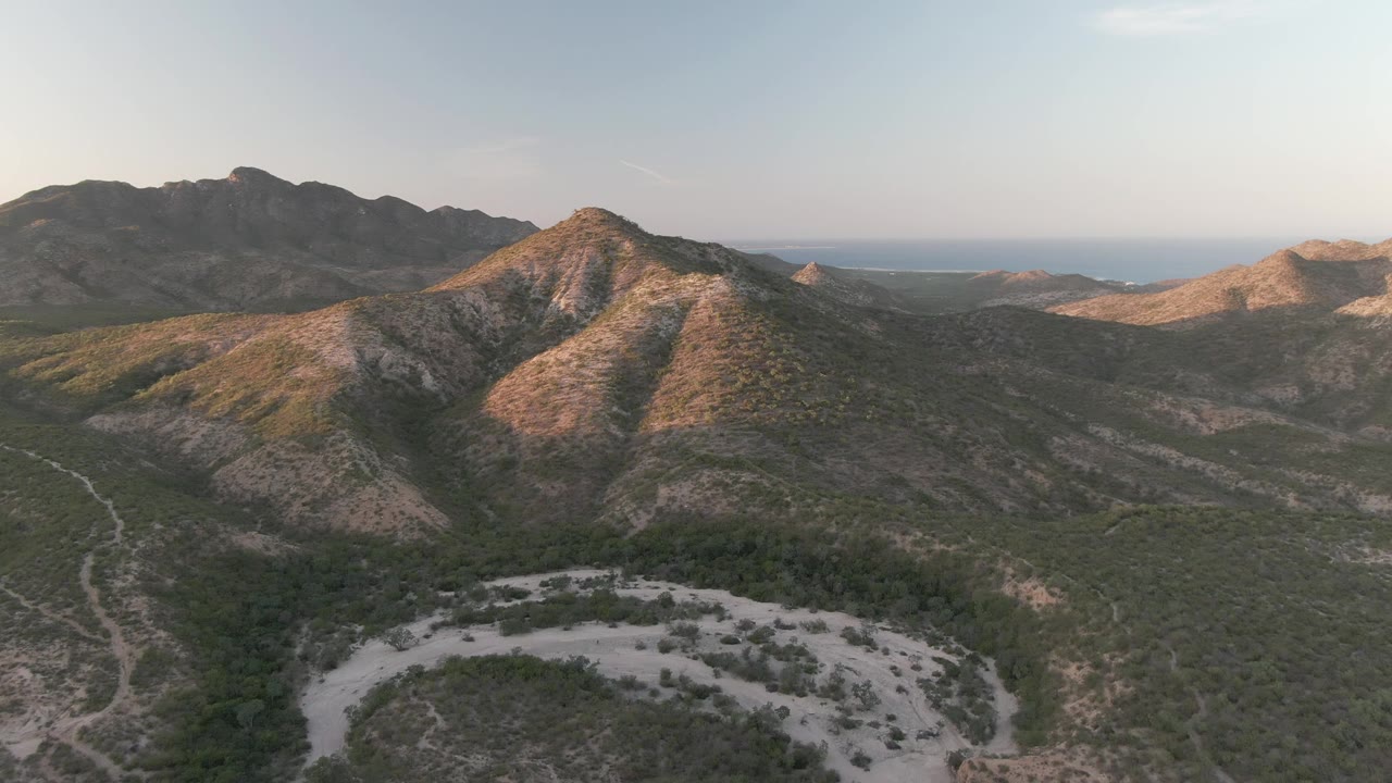 Aerial view in Cabo Pulmo, Baja California Sur, with the rounded ridge centered, darker mountains across the back, a pale track through the valley floor, distant blue water, and no visible people.