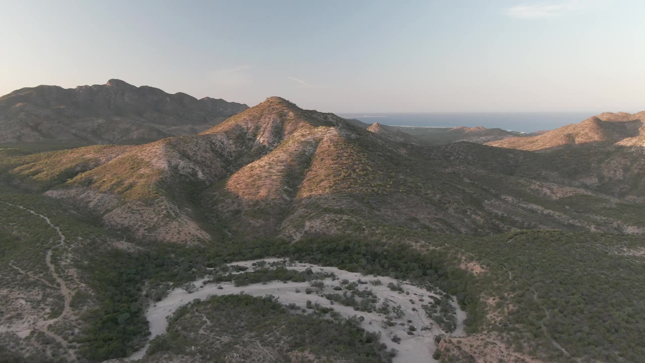Wide aerial frame from Cabo Pulmo, Baja California Sur, showing green-brown hills, a branching pale road or wash in the valley, shadowed mountains behind, faint distant water, and no visible people.