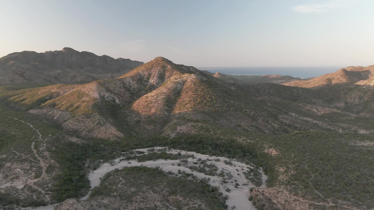 Aerial frame over Cabo Pulmo, Baja California Sur, with a broad pale wash or dirt road curving across the lower foreground, scrubland across the valley, sunlit mountain slopes, and no visible people.