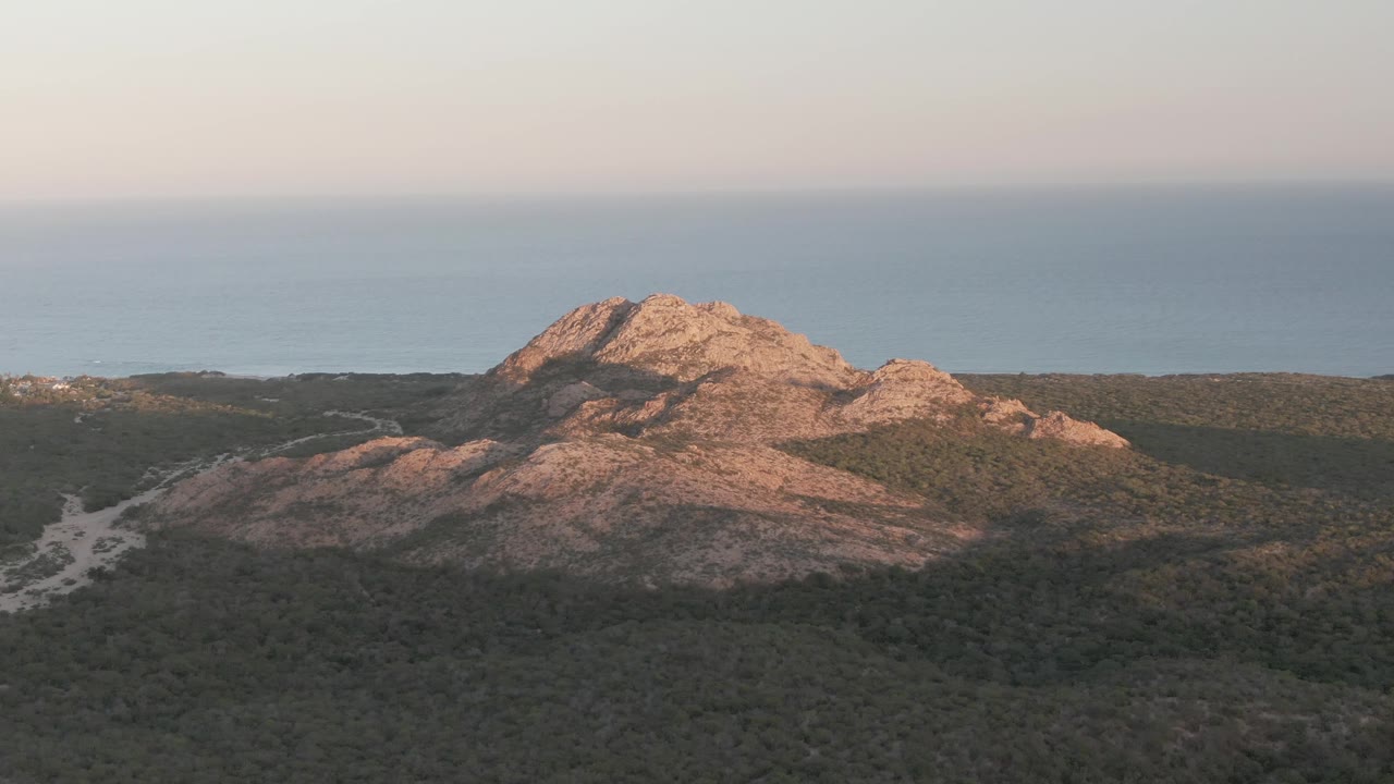 Cabo Pulmo aerial view at the start of the clip, with a tan rocky hill lit from the side, dark green scrubland below, blue ocean behind, and no visible people.