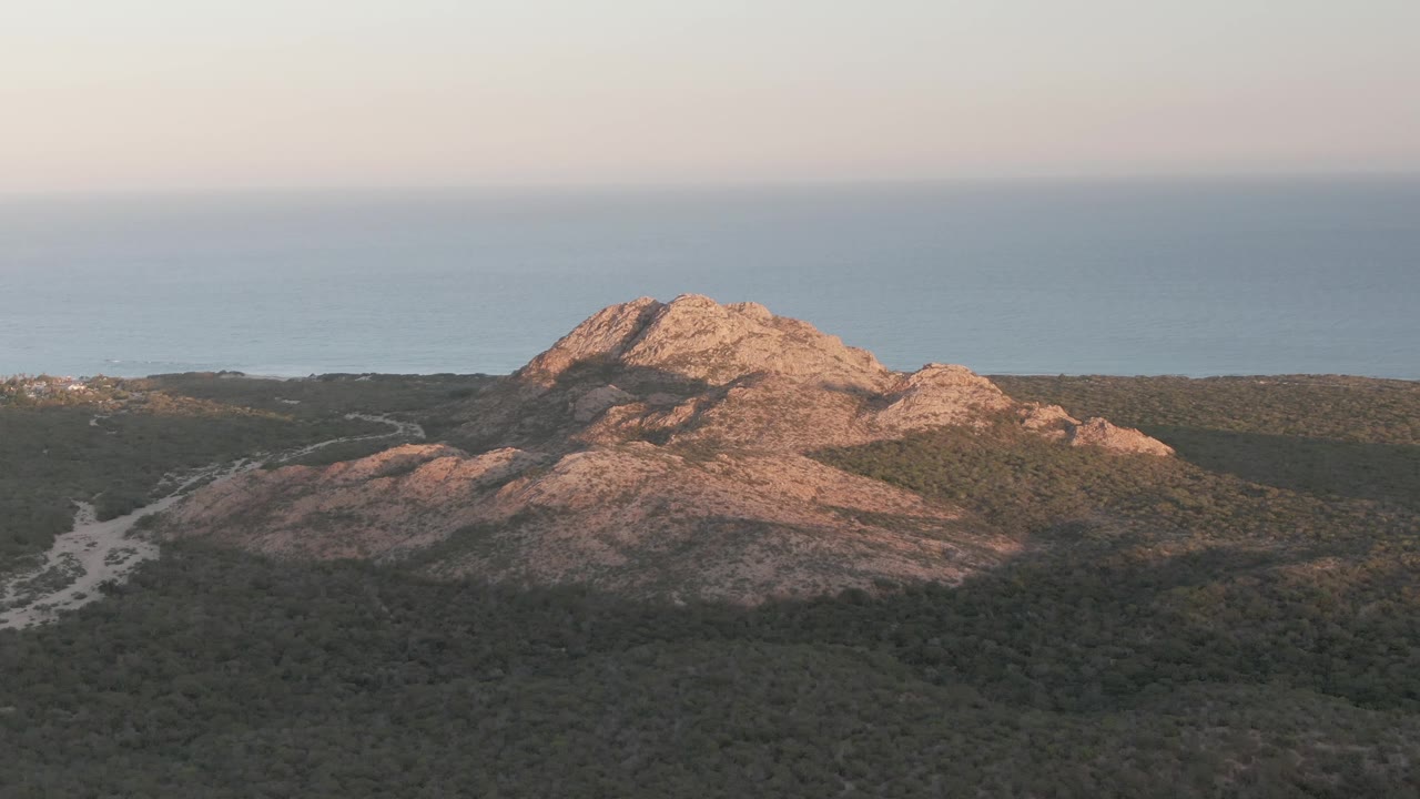 Sunlit rocky hill with scrubland and calm ocean beyond.