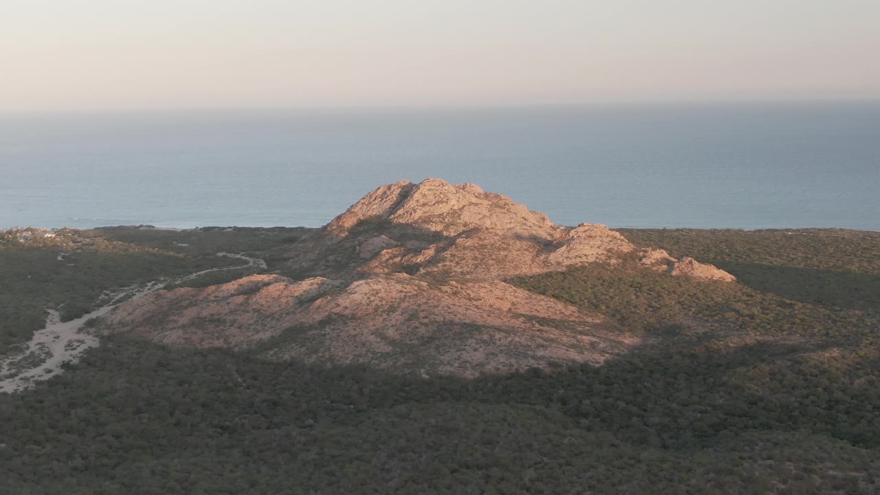 Cabo Pulmo aerial view with the hill slightly right of center, a pale track or wash visible through the scrub at left, blue water behind the ridge, and no visible people.