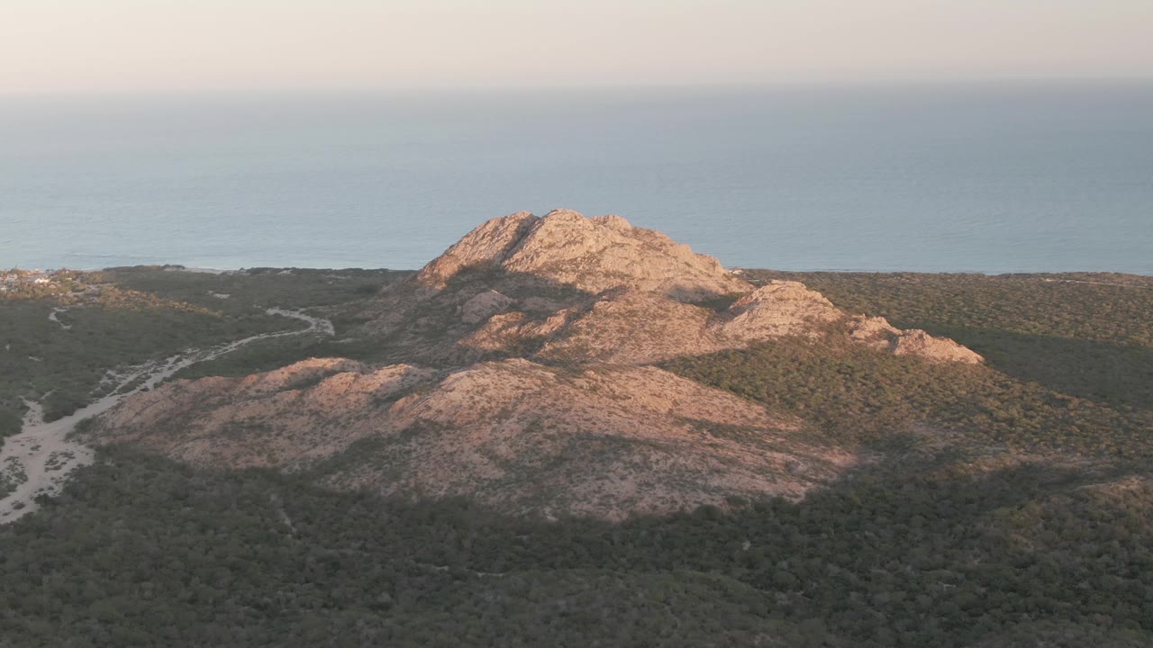 Aerial frame at 0:01 in Cabo Pulmo showing the rocky hill centered above green scrub, with a pale path threading through the lower left and ocean visible beyond, no visible people.