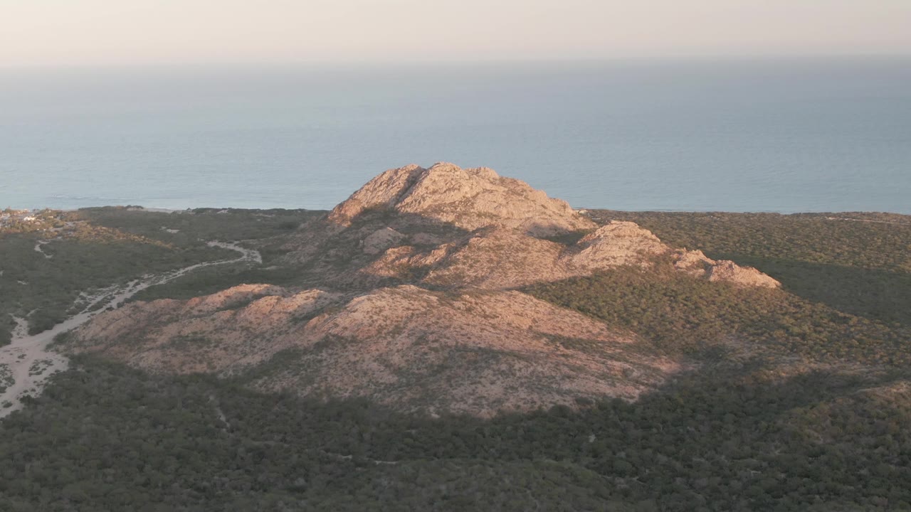 Frame at 0:02 shows warm light across the rocky Cabo Pulmo hill, darker vegetation in the foreground, a pale path below, and a soft blue ocean band in the background, no visible people.