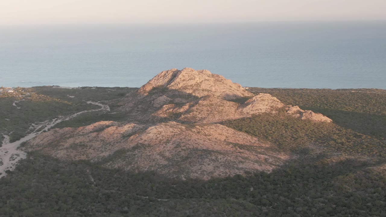 At 0:03, the Cabo Pulmo hill remains framed against the ocean, with the lower rocky slope and green vegetation showing a different balance of sun and shadow, no visible people.