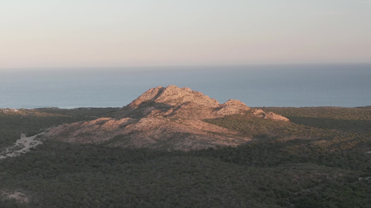 Early frame of the Cabo Pulmo hill view, showing orange-pink sunlight across the rock, darker forest in the foreground, blue ocean behind it, and no visible people.
