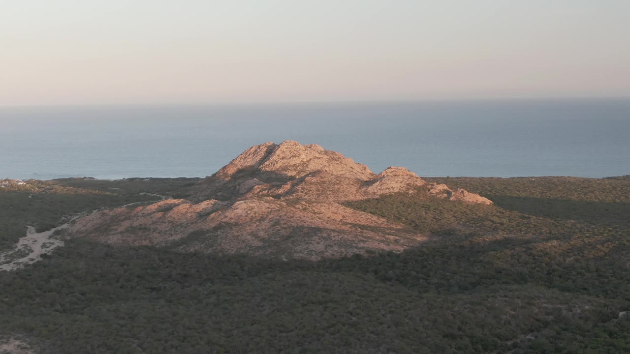 Middle frame of the Cabo Pulmo aerial view, with the rocky hill slightly left of center, dense forest below, ocean on the horizon, and no visible people.