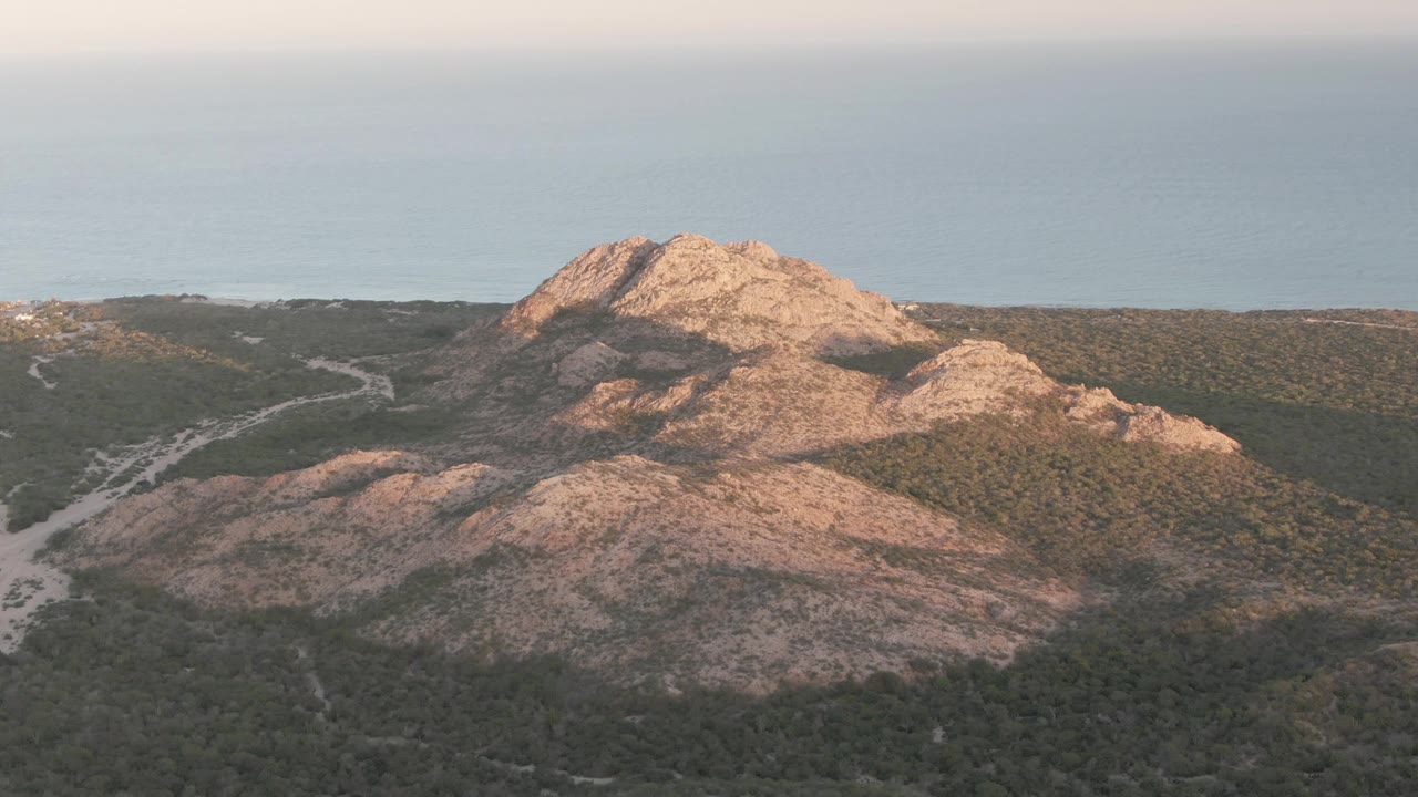 Opening aerial frame over Cabo Pulmo rocky ridges and green scrubland, with pale dirt tracks below, blue ocean in the distance, warm side light, and no visible people.