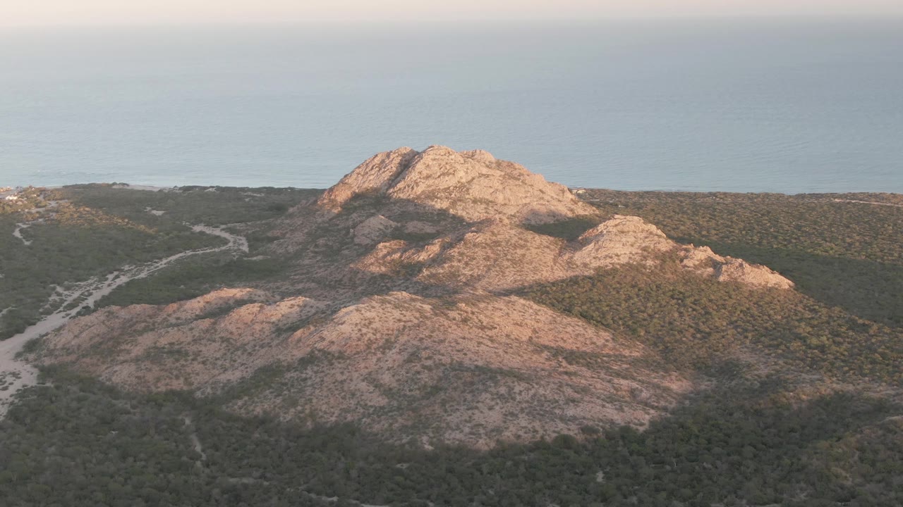 Aerial frame in Cabo Pulmo with the main tan ridge centered, dark green brush across the lower slopes, a winding dirt track to the left, ocean beyond, and no visible people.