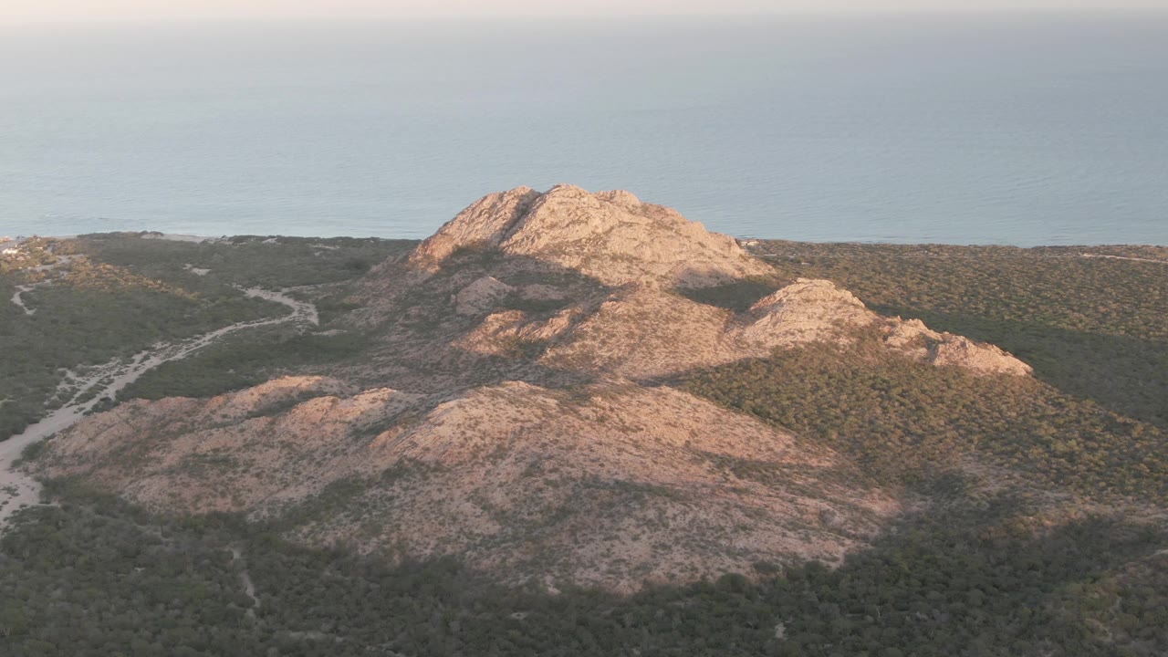 Aerial Cabo Pulmo view with sunlit tan rock formations, a shadowed lower slope, scrub-covered flats, faint dirt roads, blue ocean background, and no visible people.