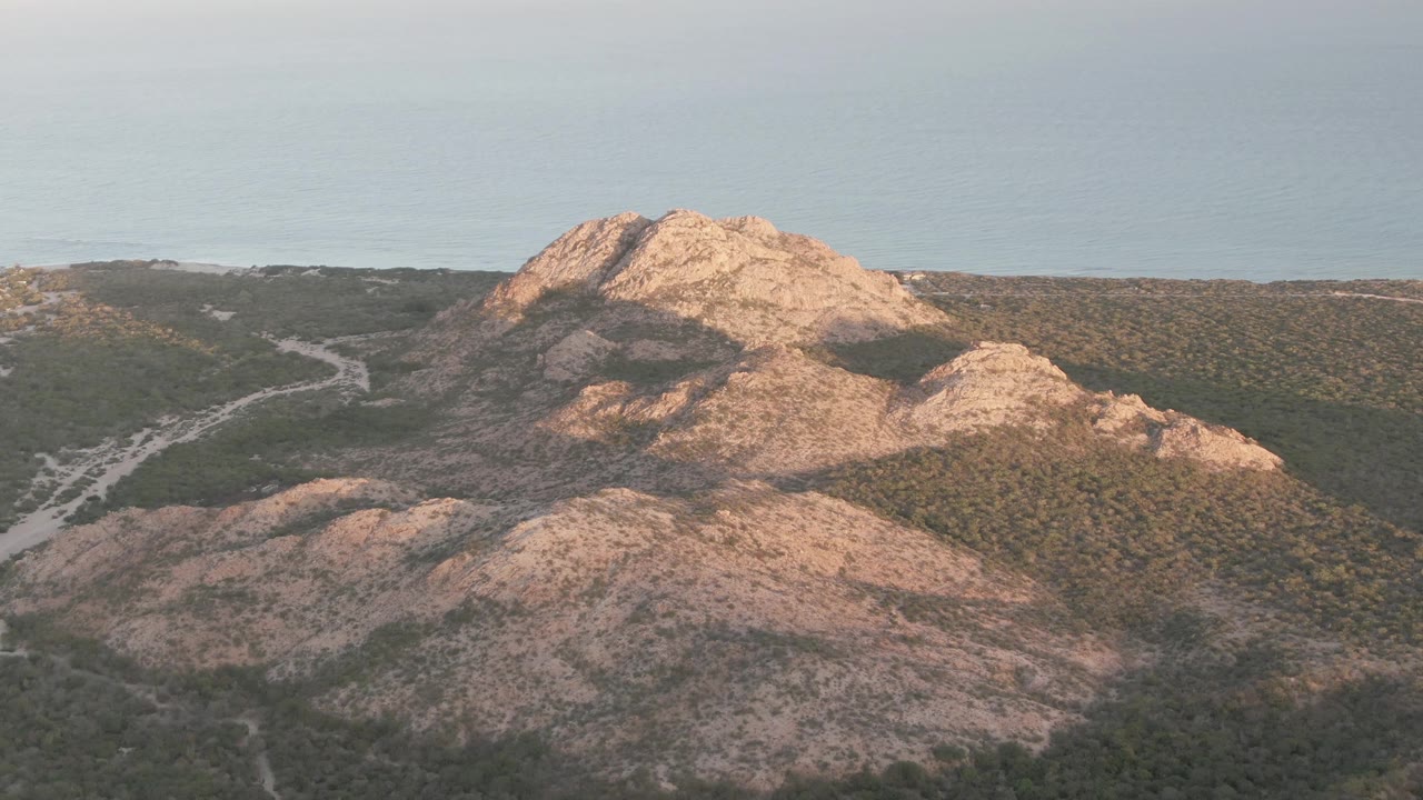 Opening frame of Cabo Pulmo rocky hills with green scrub, a pale winding road at left, ocean beyond the ridge, and no visible people.