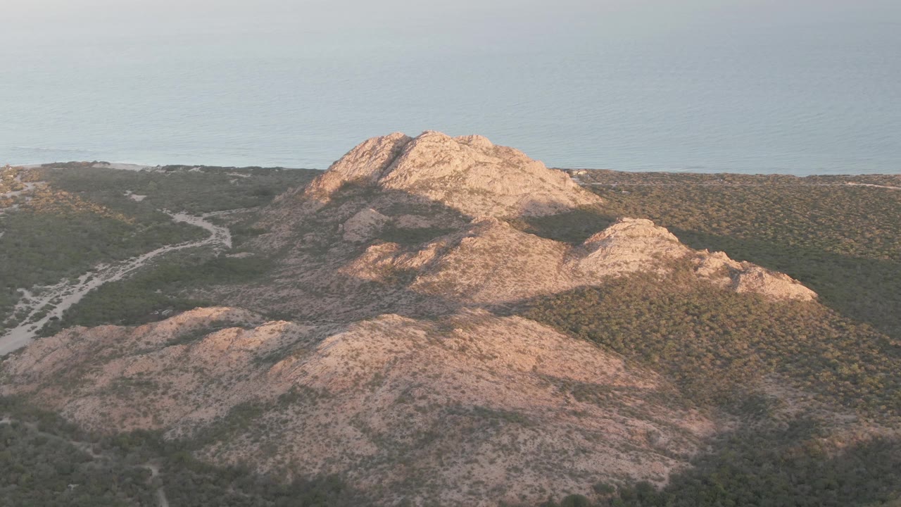 Cabo Pulmo aerial frame showing sunlit tan ridges, darker scrub-covered slopes, a winding road through the midground, ocean in back, and no visible people.
