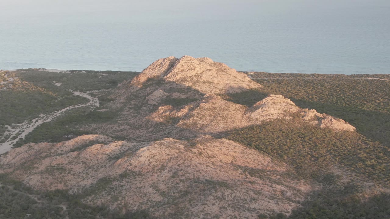 Cabo Pulmo frame with the main rocky hilltop centered, scrubland spreading across the slopes, road bends visible below, ocean behind, and no visible people.