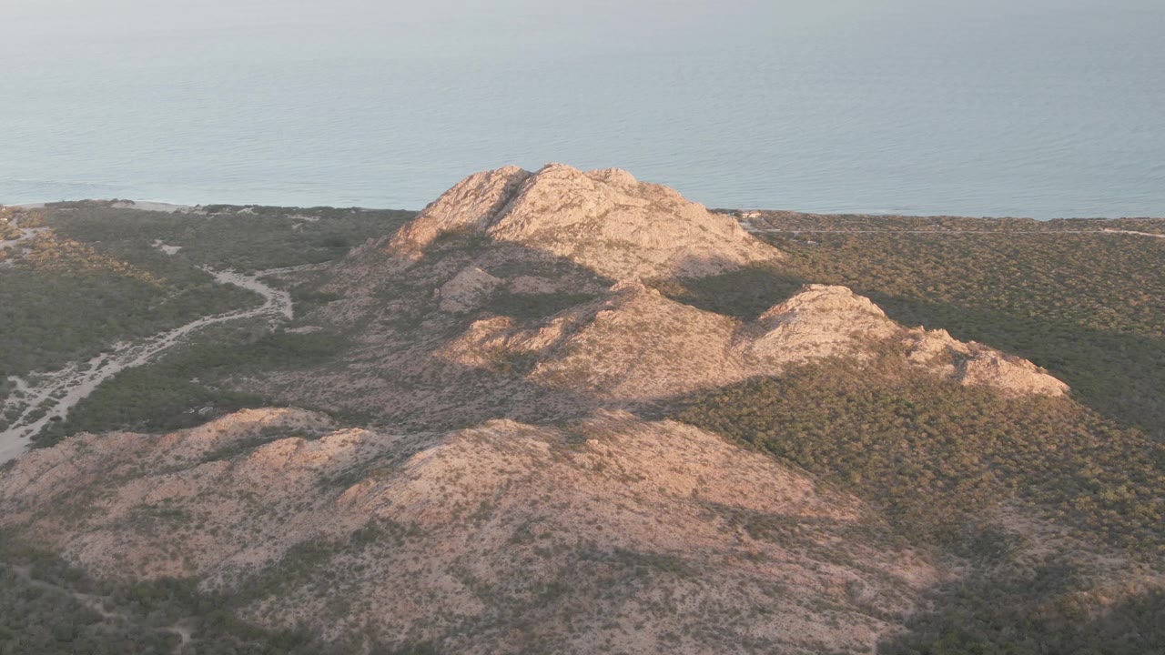 Later frame from Cabo Pulmo showing a rocky foreground slope, the winding road along the left side, dense green scrub, ocean beyond the ridge, and no visible people.
