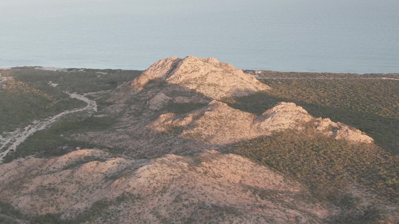 Aerial frame over Cabo Pulmo, Baja California Sur, showing tan rocky hills, green scrub, a pale winding road in the valley, distant blue horizon, and no visible people.