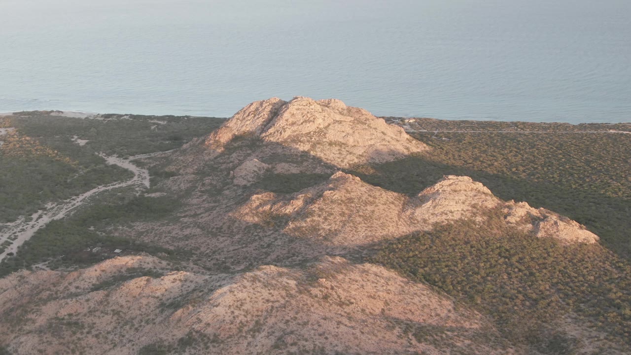 Cabo Pulmo aerial frame showing the road bending through green hillside scrub, tan rocky peaks in the foreground and midground, a pale blue distance, and no visible people.