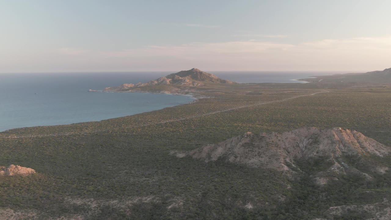 Aerial view over Cabo Pulmo, Baja California Sur, with rocky foreground hills, green scrubland, a pale road, blue ocean on the left, distant mountains, and no visible people.