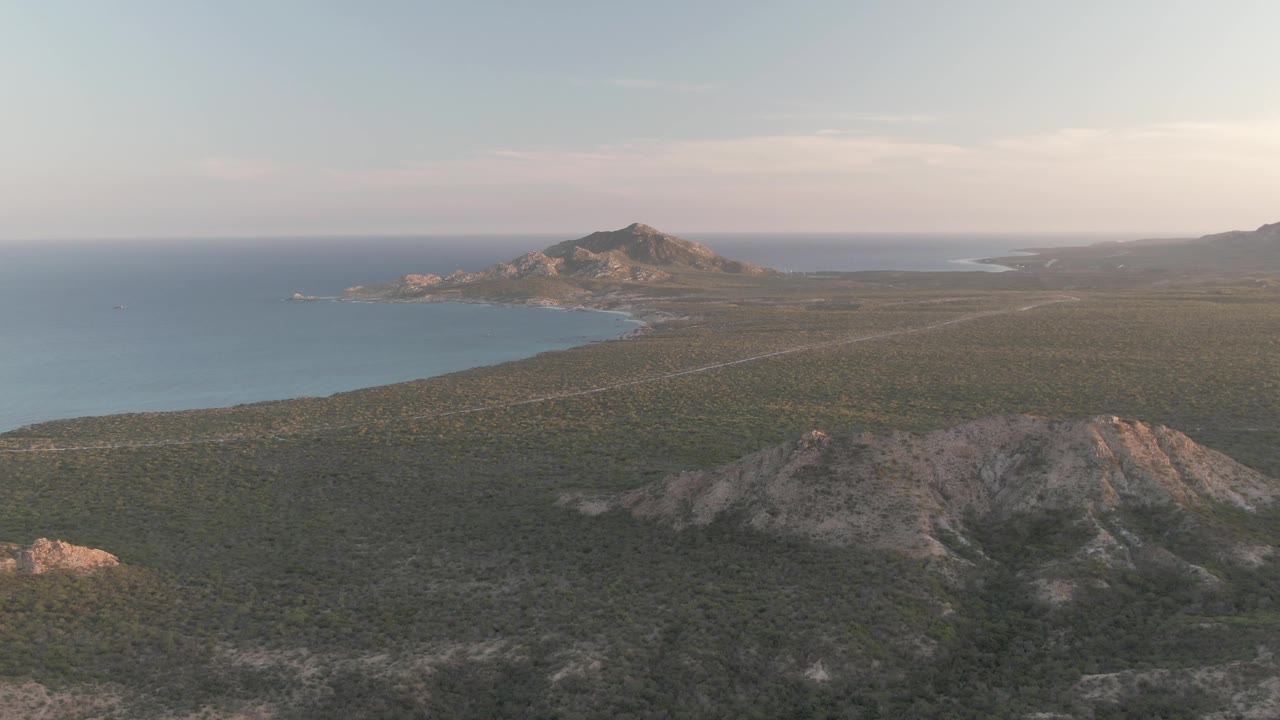 Aerial Cabo Pulmo view with the road stretched across green terrain, ocean and shoreline at left, a rocky hill near the foreground, distant mountains, soft daylight, and no visible people.