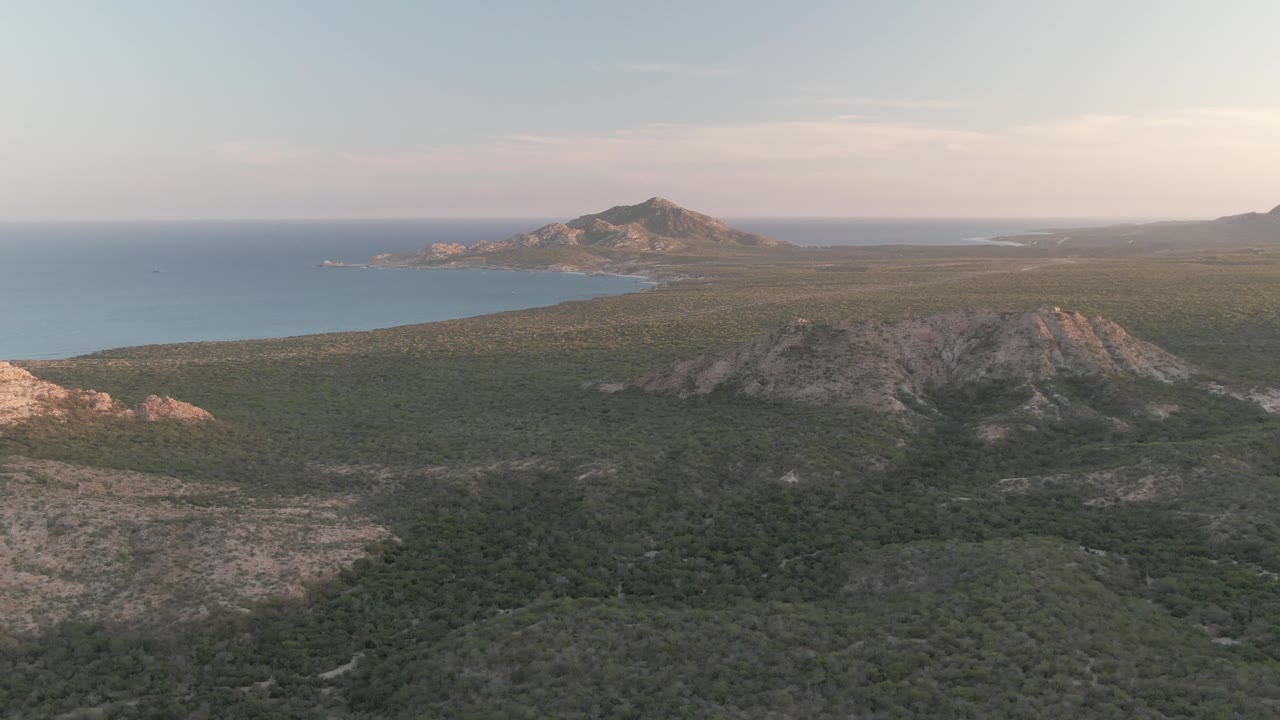 Cabo Pulmo aerial frame showing scrub-covered terrain in the foreground, rocky ridges in the center, blue bay water to the left, and no visible people.