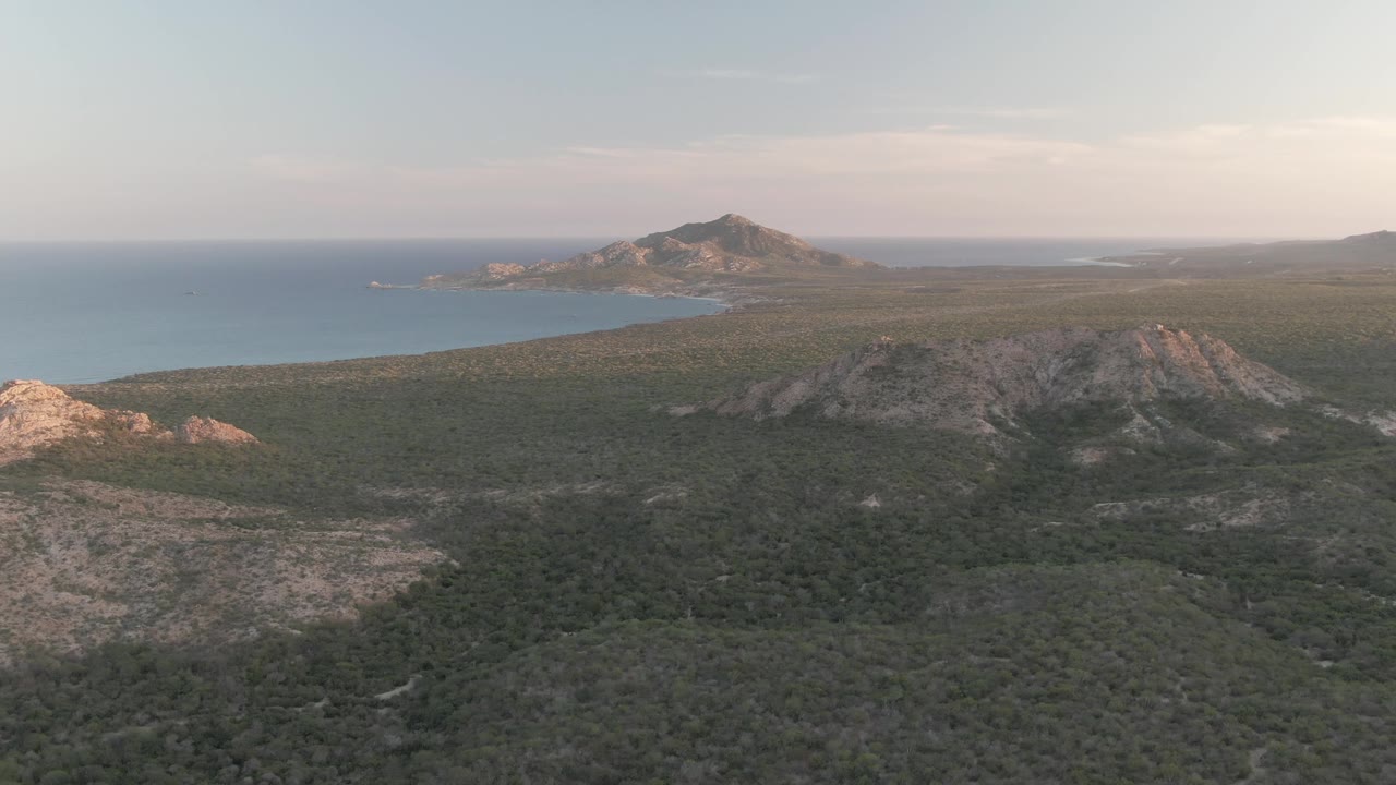 Cabo Pulmo dusk frame with rocky outcrops at left, scrubland across the foreground, central hills, blue bay water in back, and no visible people.