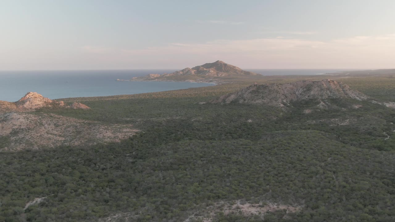 Early aerial moment in Cabo Pulmo showing scrubby green terrain in the foreground, sunlit rock on the left, darker ridges to the right, ocean in back, and no visible people.