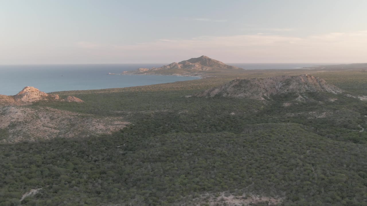 Mid-pan aerial frame over Cabo Pulmo with the rocky central ridge more prominent, green brush filling the lower frame, blue water along the left background, and no visible people.