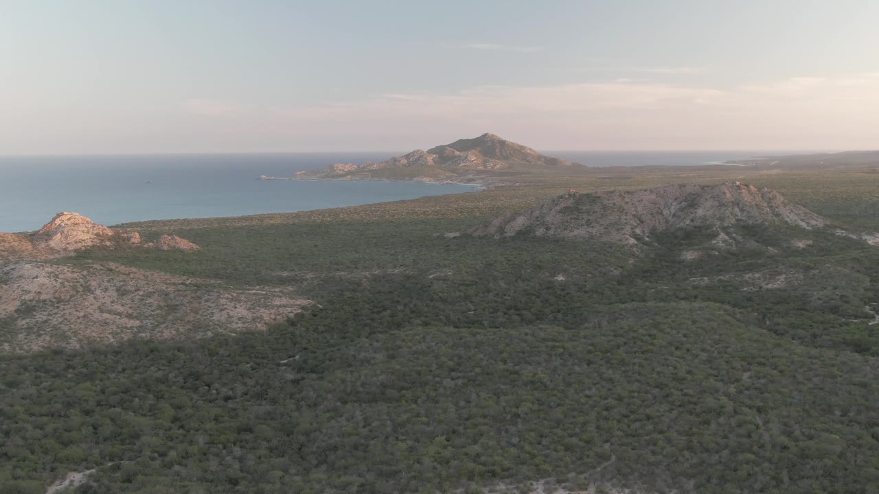 Later aerial frame in Cabo Pulmo showing more foreground vegetation, a pale rocky slope at lower left, rounded hills across the middle, blue ocean behind, and no visible people.