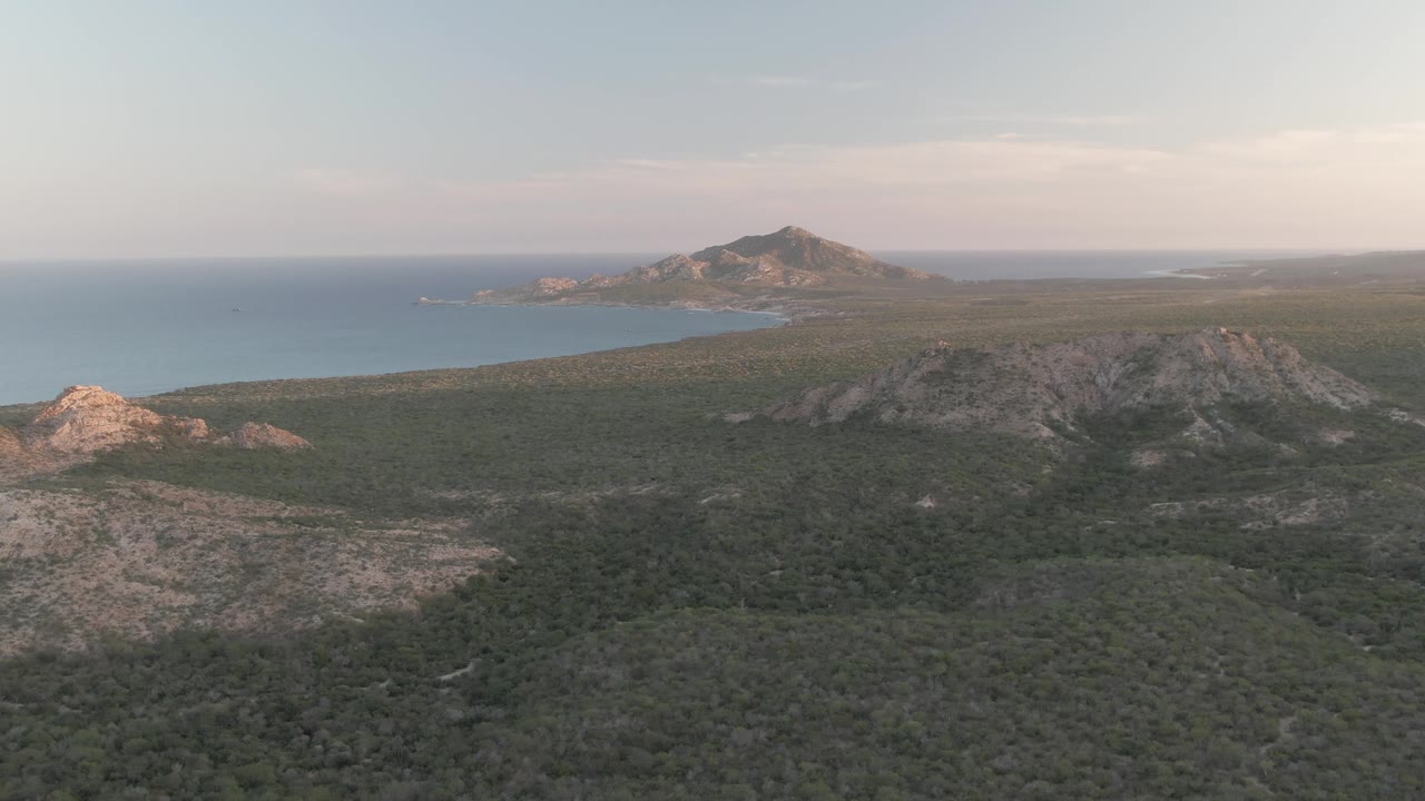 Aerial frame in Cabo Pulmo with broad green terrain in the foreground, rocky ridges in the midground, a blue ocean edge to the left, distant hills, and no visible people.