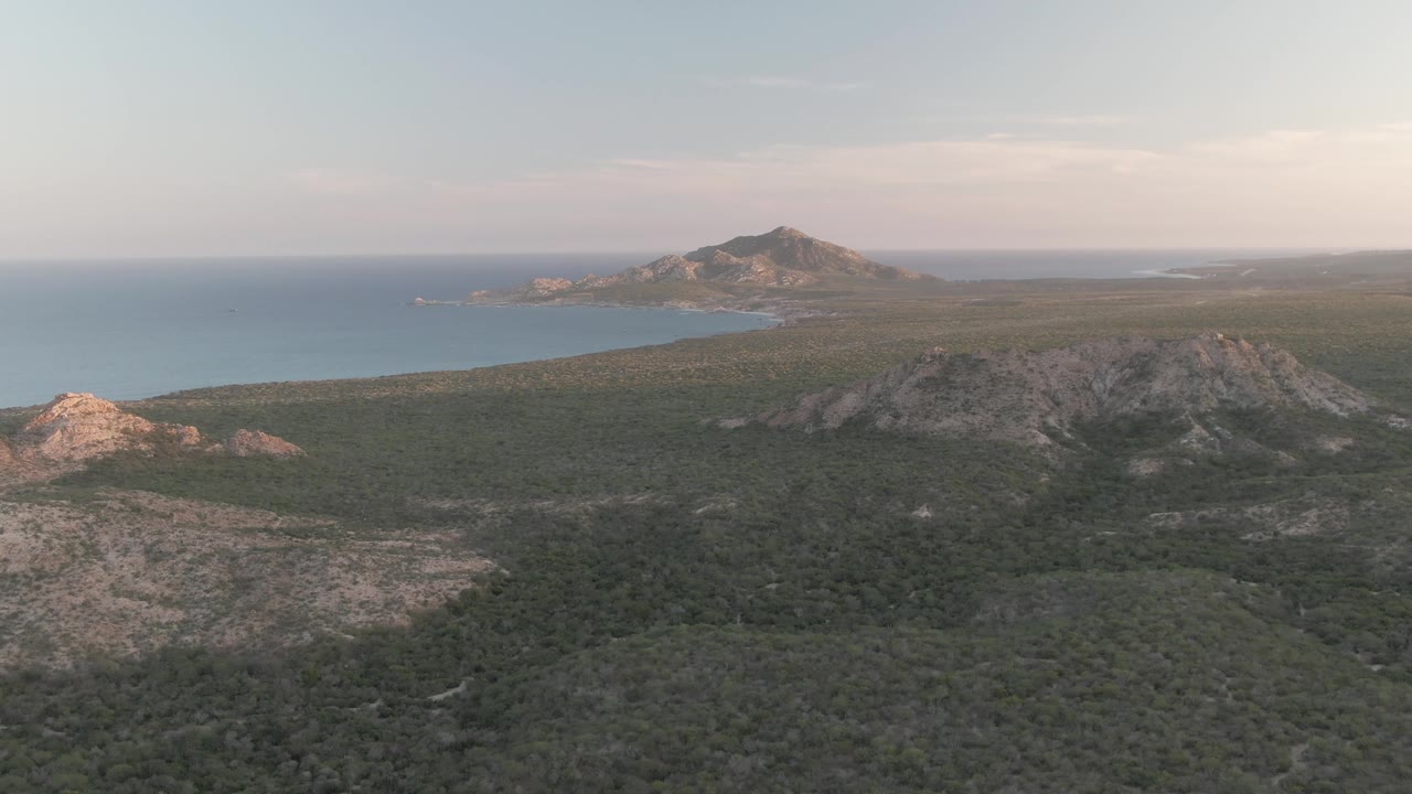 Aerial frame over Cabo Pulmo showing sunlit rocky outcrops at left, a scrub-covered plain, a blue bay, a distant headland near the horizon, and no visible people.