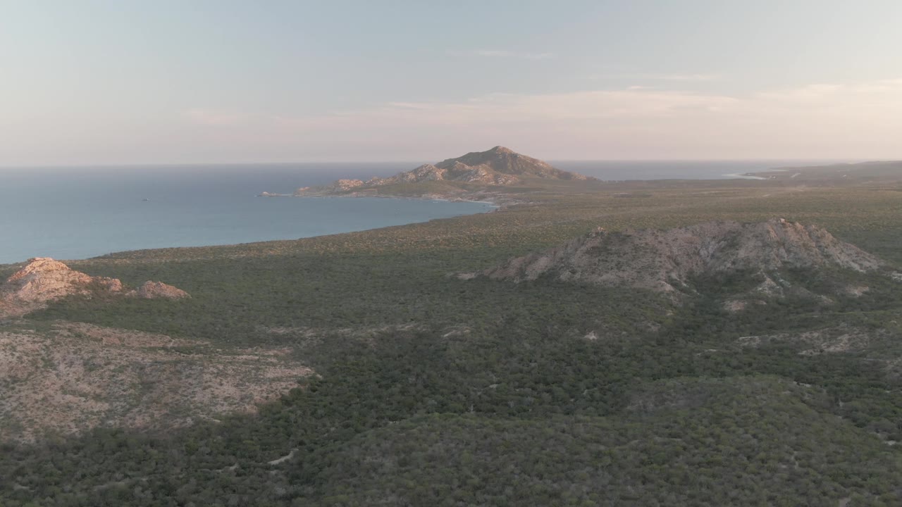 Aerial frame in Cabo Pulmo with a rocky hill in the lower right, green vegetation across the valley, blue ocean to the left, distant mountain shapes, and no visible people.