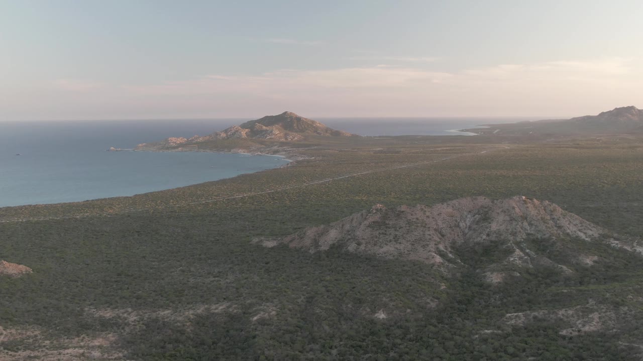 Cabo Pulmo aerial frame with the ocean spread across the left side, rocky green foreground terrain below, the low landform near the shore, distant hills on the horizon, and no visible people.
