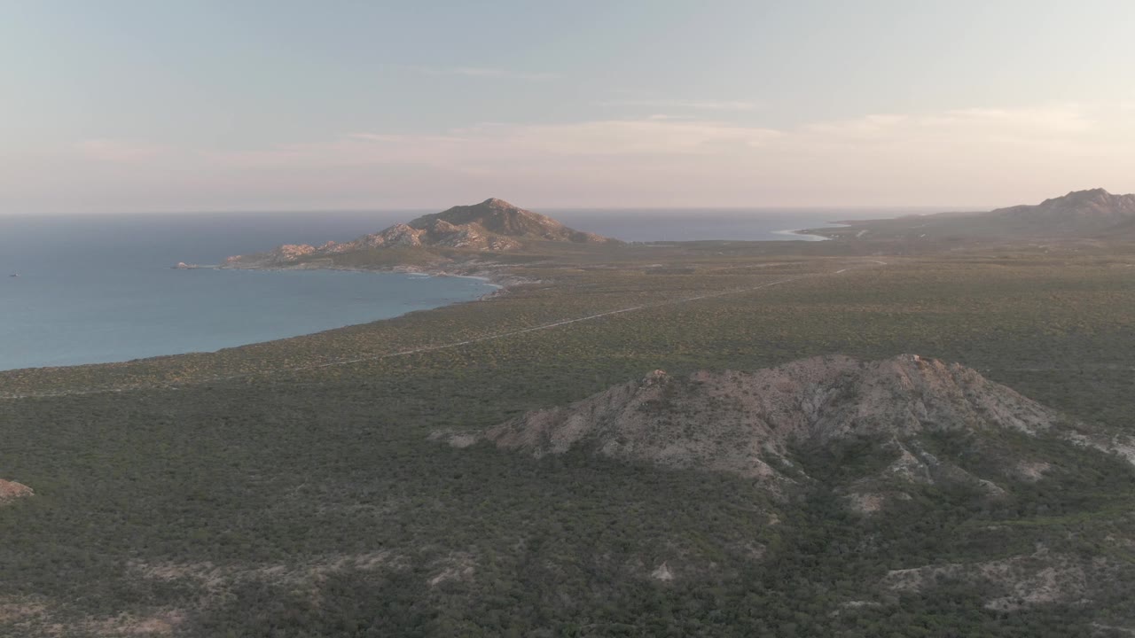 Cabo Pulmo view with the rocky foreground ridge lower in frame, the bay and shoreline visible at left, broad green terrain across the center, distant hills at right, soft sky, and no visible people.