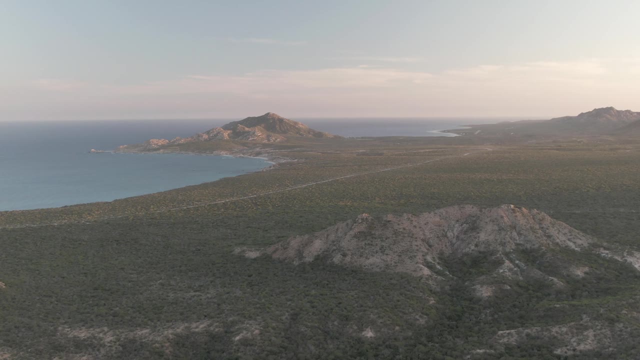 Mid-clip Cabo Pulmo frame with the shoreline and blue water more prominent on the left, rocky scrubland filling the lower half, distant hills set under a pale sky, and no visible people.