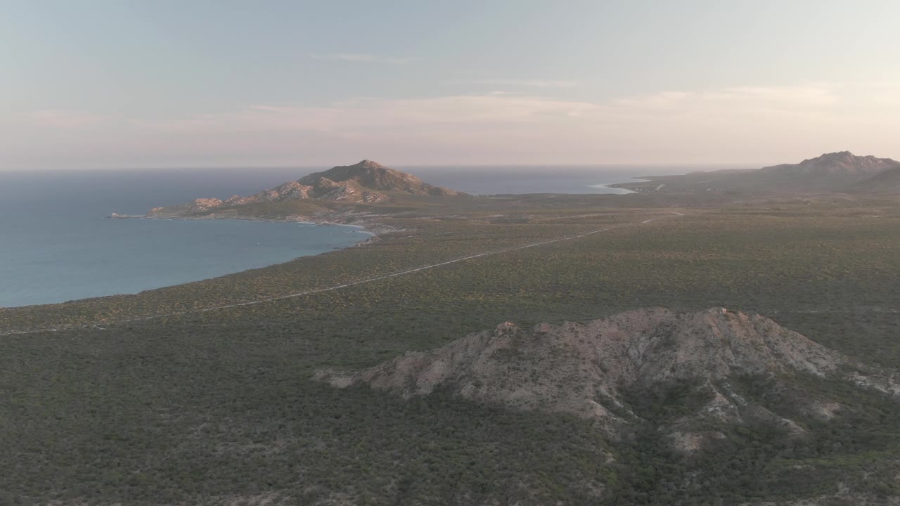 Wide aerial frame over Cabo Pulmo with a rocky foreground ridge, a pale road across dry terrain, blue ocean at left, distant hills, soft low light, and no visible people.
