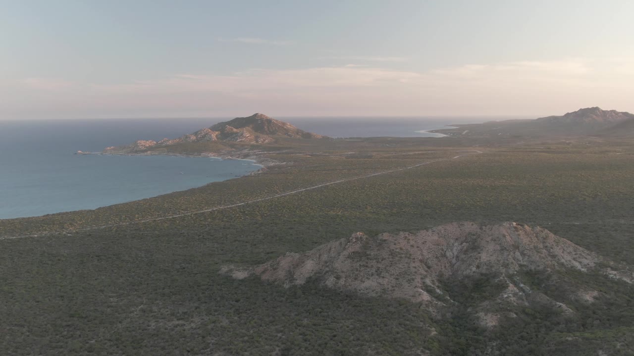 Second frame of the Cabo Pulmo aerial view, with the road running diagonally through scrubland, ocean water along the left shoreline, rocky ridges below, distant hills, and no visible people.