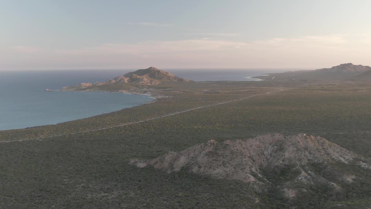 Third frame shows Cabo Pulmo from a high aerial angle, with the road line crossing the dry plain, a rocky ridge in the foreground, ocean and shoreline to the left, hazy mountains in the distance, and no visible people.