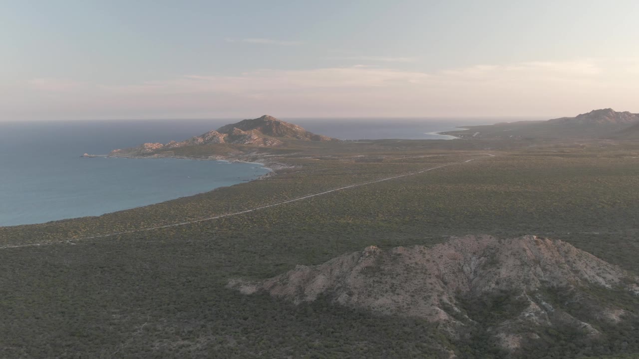 Wide drone view of ocean, shoreline, road, and rocky desert terrain.