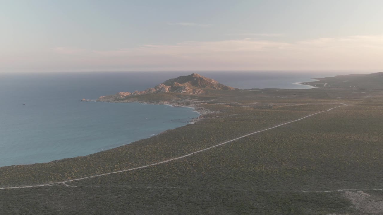 Aerial frame over Cabo Pulmo showing the road angled across dark scrubland, the ocean and shoreline on the left, rocky hills near center, and no visible people.