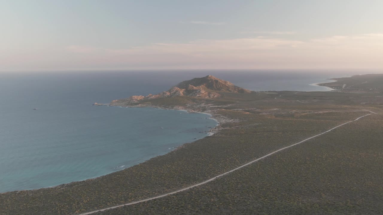 Aerial coastline with blue ocean, coastal road, scrub terrain, rocky hill, and small boat offshore.