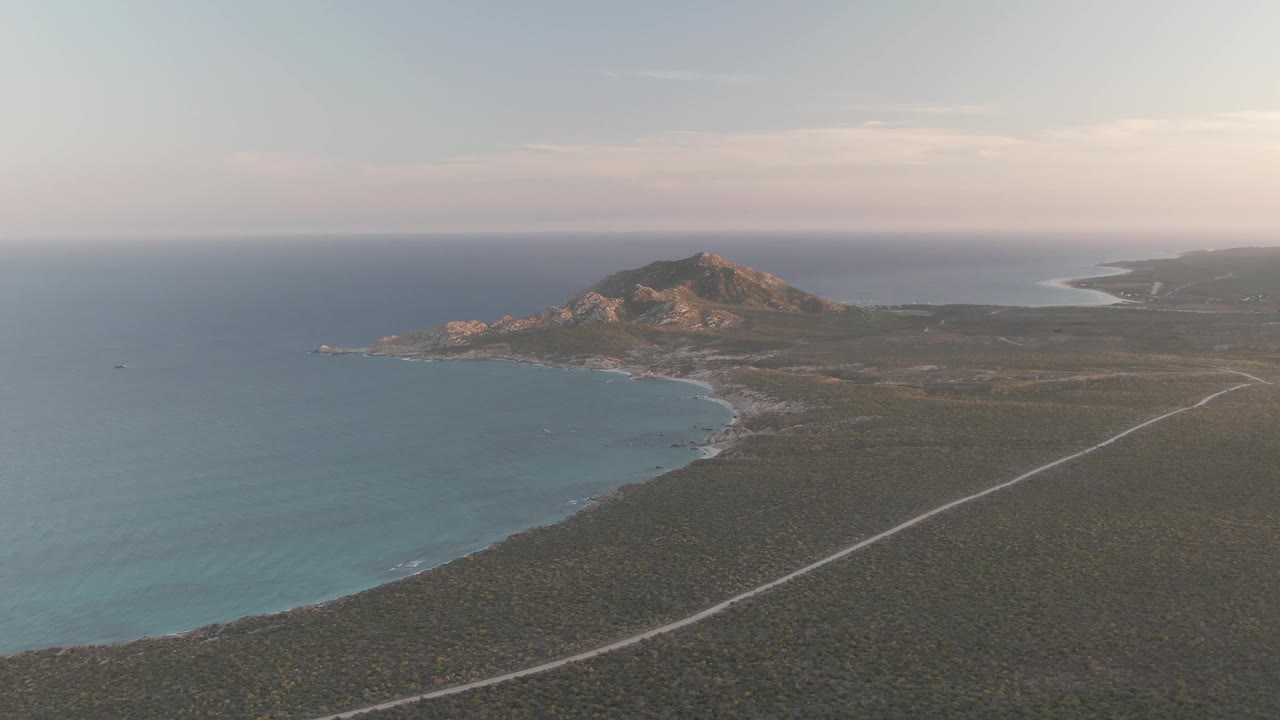 Aerial view over Cabo Pulmo with the curved bay filling more of the left side, the diagonal coastal road crossing the scrubland, distant rocky hills, soft horizon clouds, and no visible people.
